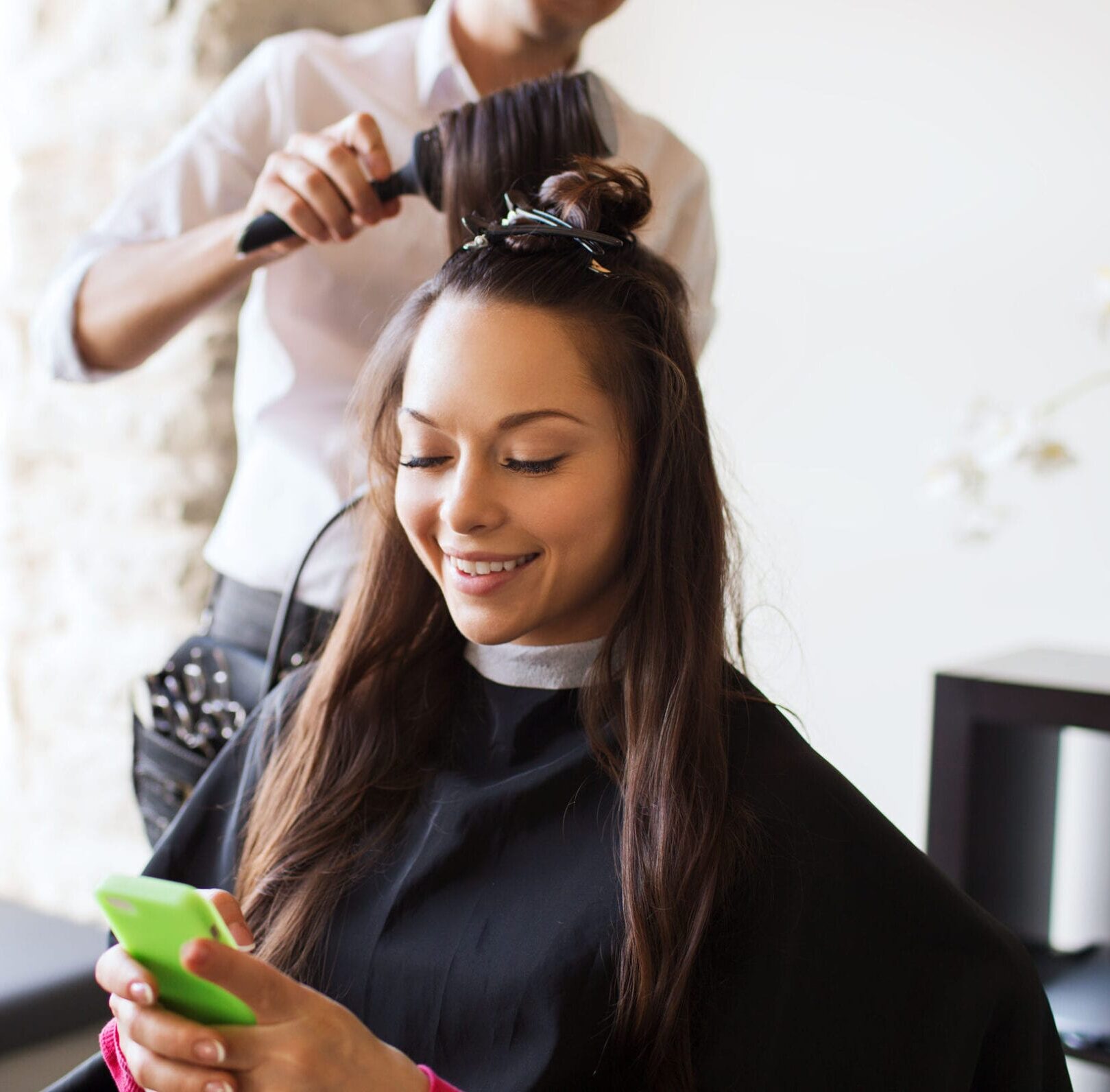 happy young woman with smartphone and hairdresser making hair styling at salon